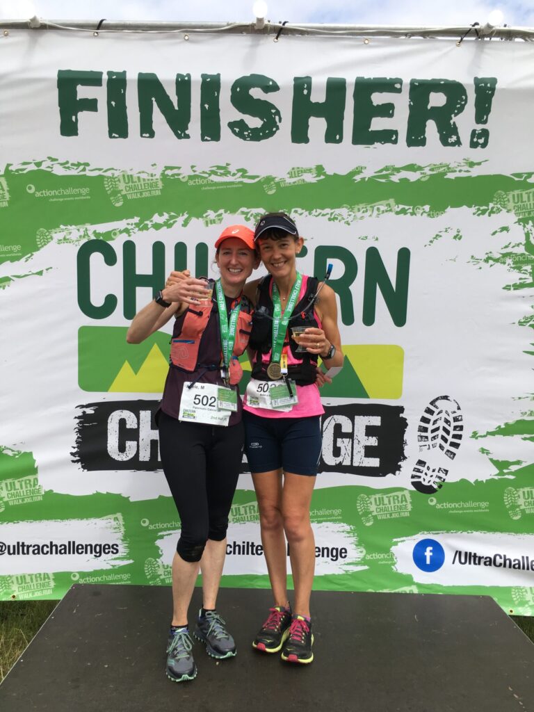 Catherine in running outfit with another runner in front of a Finisher poster